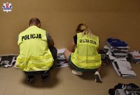 odwrócony tyłem policjant i funkcjonariusza Urzędu Celnego, na podłodze leżą zabezpieczone ubrania